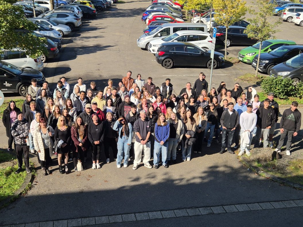 Große Gruppe von Menschen steht auf einem Parkplatz, im Hintergrund mehrere geparkte Autos und Bäume.