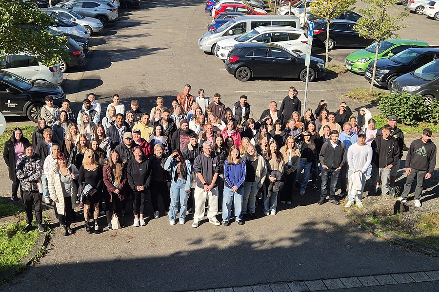 Große Gruppe von Menschen steht auf einem Parkplatz, im Hintergrund mehrere geparkte Autos und Bäume.