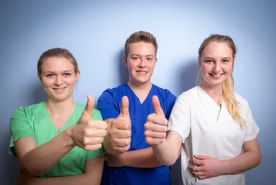 Drei Personen in medizinischer Kleidung zeigen Daumen hoch. Links trägt eine Person ein grünes Oberteil, in der Mitte ein blaues, rechts ein weißes.