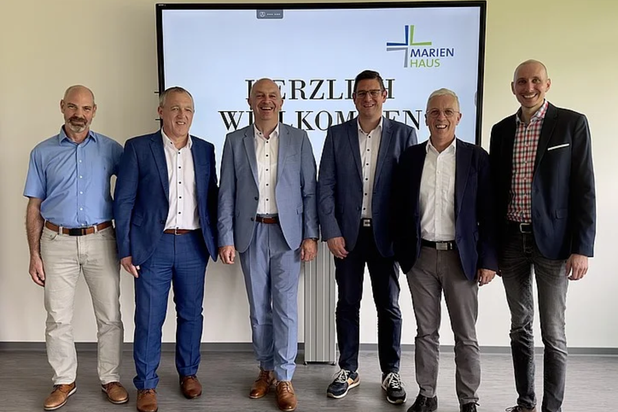 Sechs Männer in Anzügen und Hemden stehen vor einem Bildschirm mit dem Text 'HERZLICH WILLKOMMEN' und dem Logo von 'MARIENHAUS'.