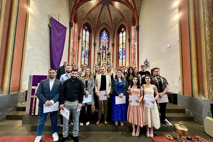 Gruppe von Personen in formeller Kleidung, die in einer Kirche stehen. Im Hintergrund sind bunte Fenster und religiöse Statuen sichtbar. Einige halten Dokumente.