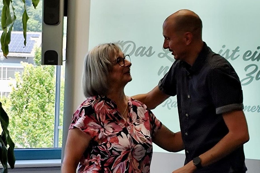 Eine Frau mit grauen Haaren trägt ein blühendes Oberteil und steht neben einem Mann in einem dunklen Hemd.