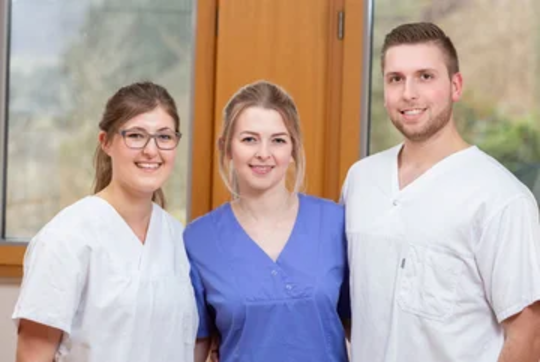 Drei Personen in medizinischer Kleidung stehen nebeneinander. Links trägt eine Person ein weißes Oberteil, in der Mitte ein blaues Oberteil und rechts ein weiteres weißes Oberteil.