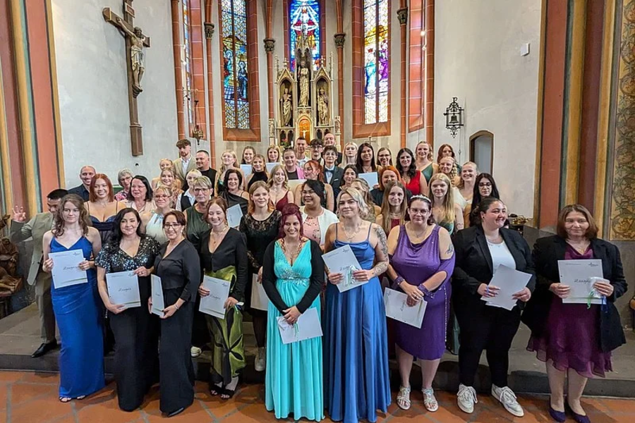 Gruppenfoto von 24 Personen in formeller Kleidung, die Urkunden halten, in einer Kirche mit bunten Glasfenstern im Hintergrund.