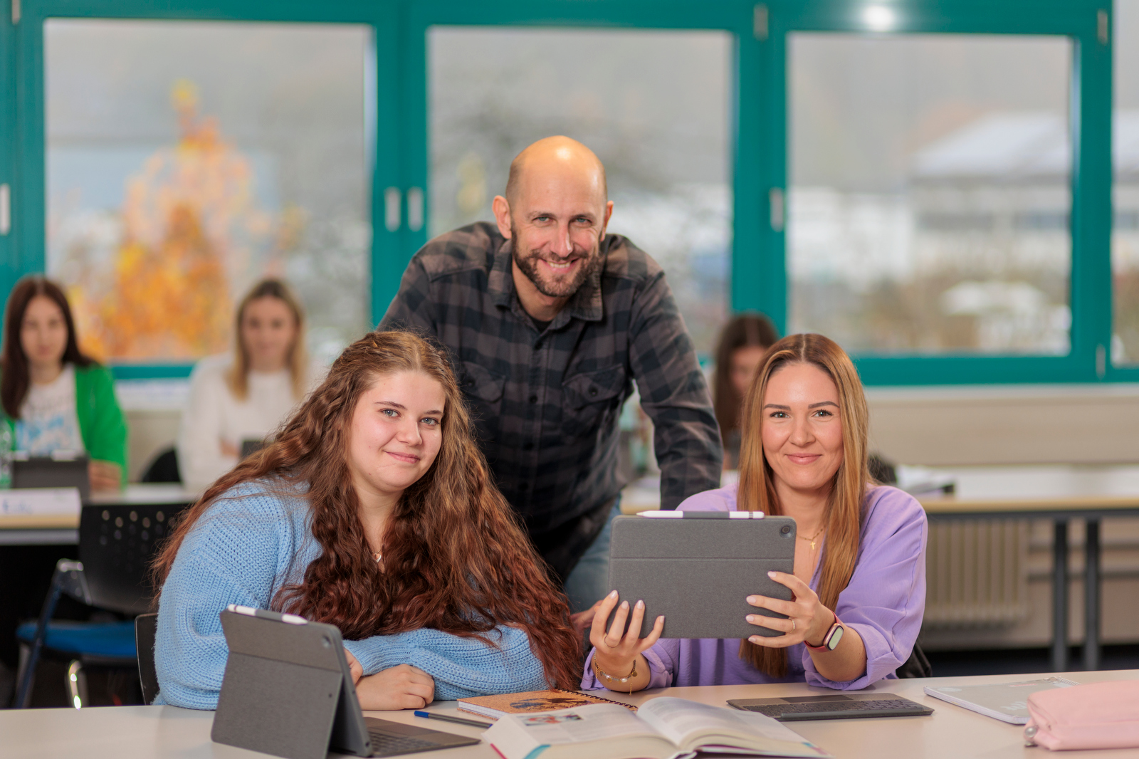 Drei Personen sitzen an einem Tisch mit Tablets und einem aufgeschlagenen Buch in einem Klassenraum mit großen Fenstern im Hintergrund