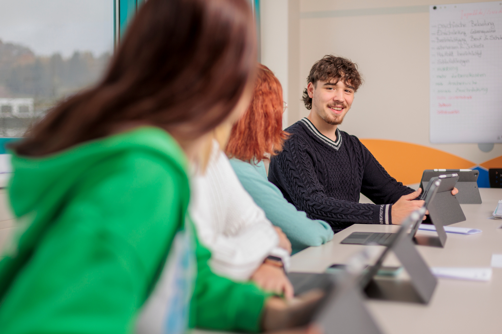 Drei Personen sitzen an einem Tisch mit Tablets und Laptops, eine Person spricht und gestikuliert, im Hintergrund eine Pinnwand mit Notizen.