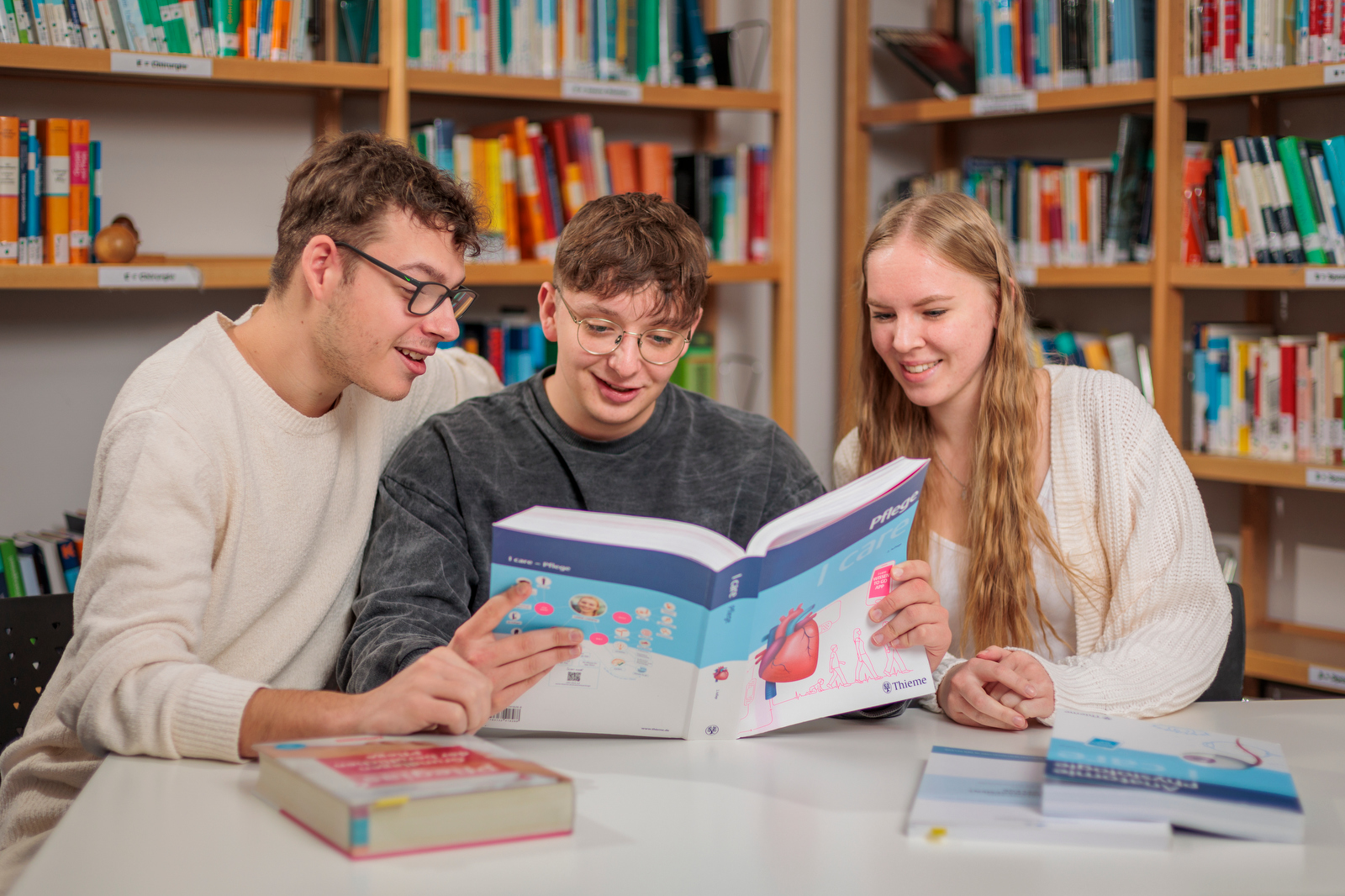 Drei Personen sitzen an einem Tisch in einer Bibliothek und lesen gemeinsam ein Buch mit anatomischen Abbildungen