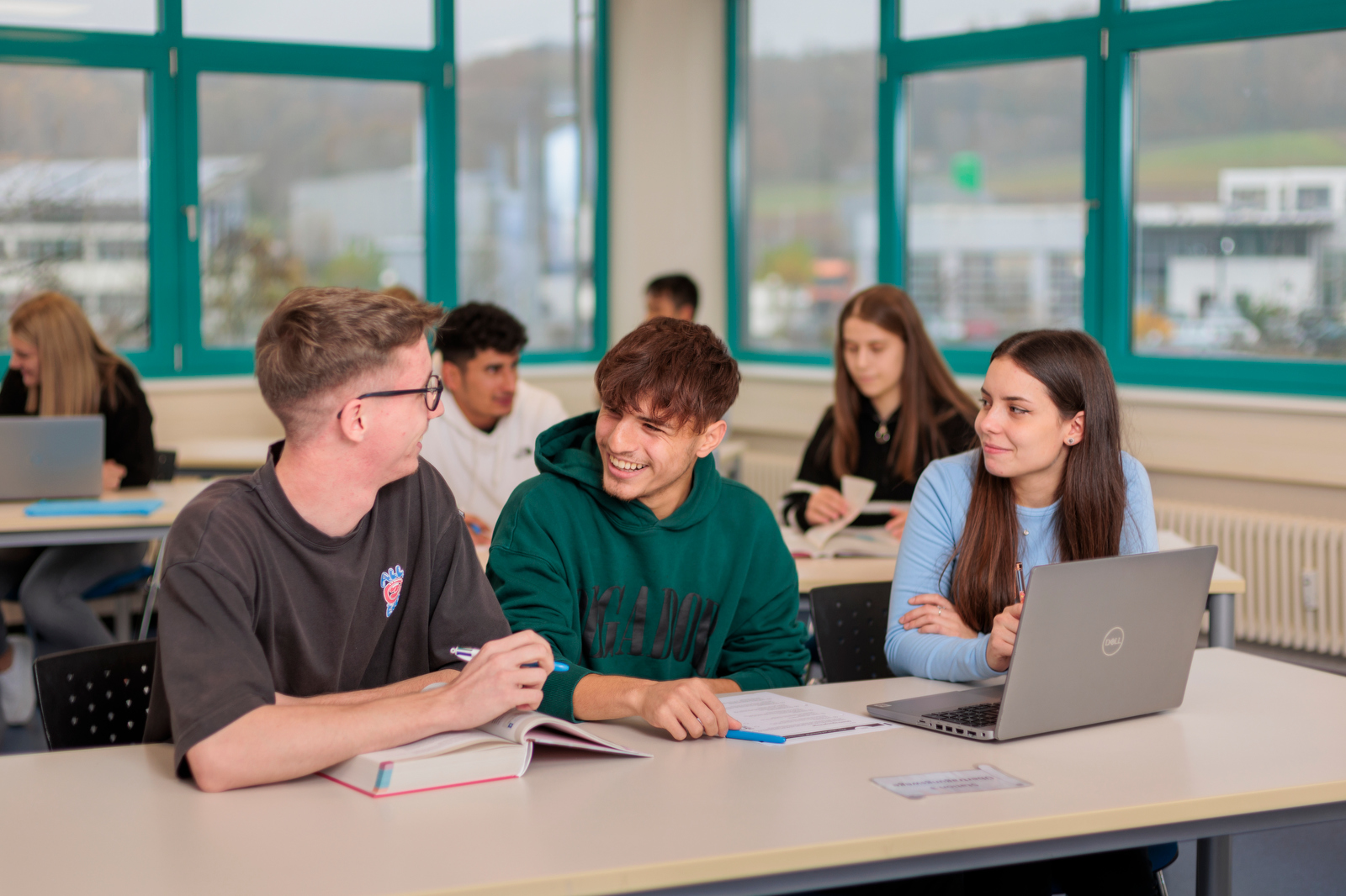 Gruppe von Jugendlichen sitzt an Tischen in einem Klassenraum mit großen Fenstern und arbeitet mit Büchern und Laptop