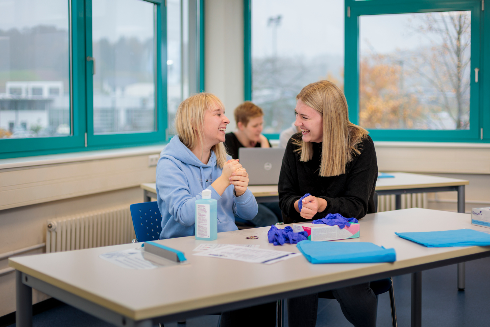 Zwei Personen sitzen an einem Tisch mit Schreibtischutensilien und unterhalten sich in einem hellen Raum mit großen Fenstern.