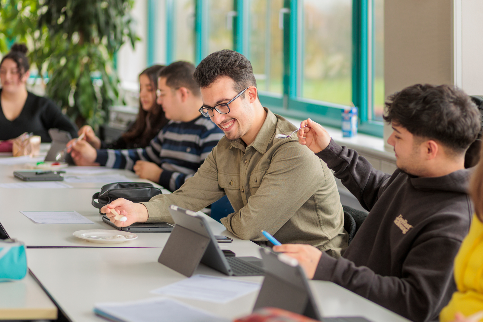 Mehrere Personen sitzen an einem Tisch mit Tablets, Notizbüchern und Stiften in einem hellen Raum mit großen Fenstern.