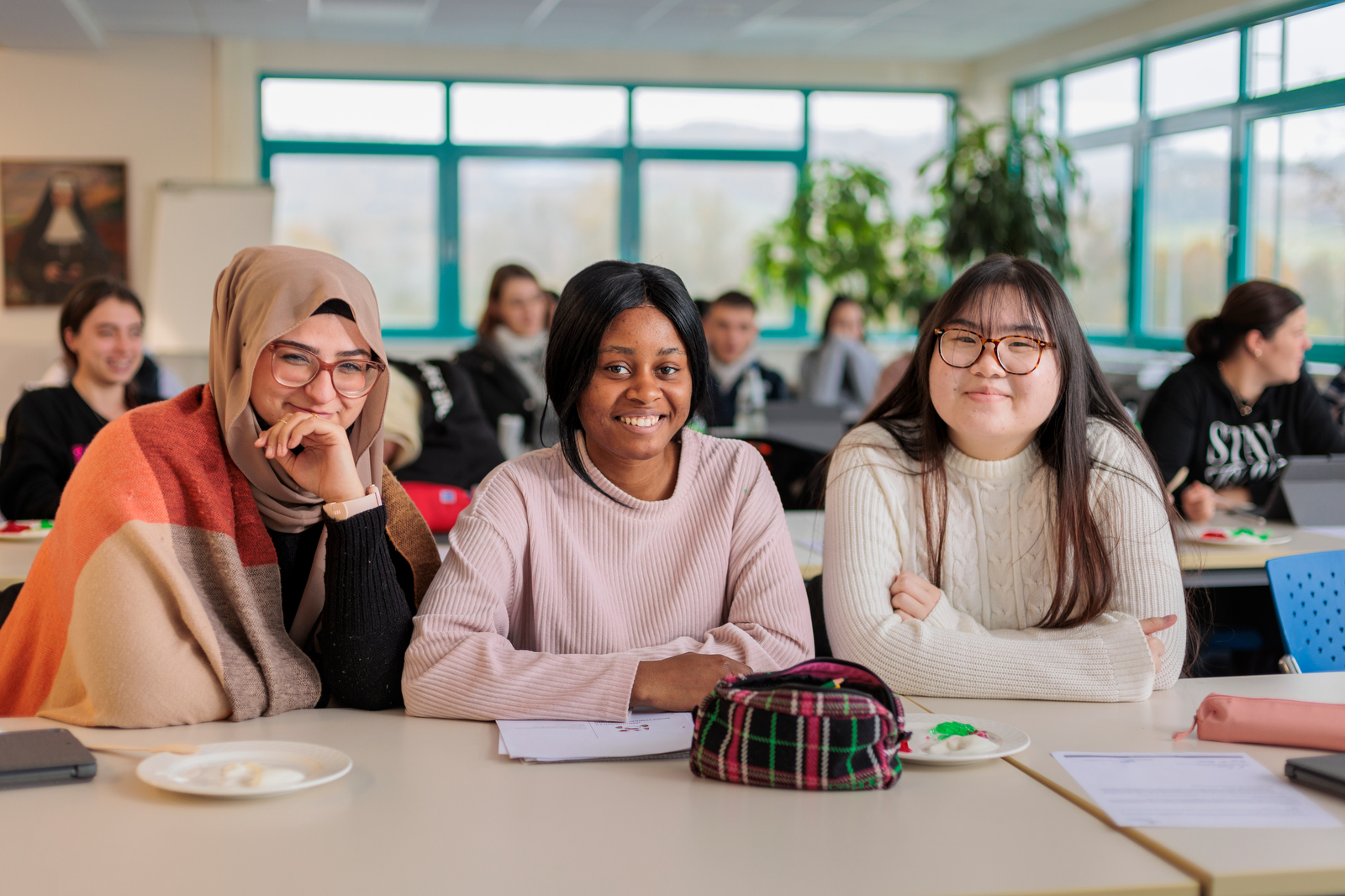 Drei junge Frauen sitzen an einem Tisch in einem Klassenraum mit Notizbüchern und Stiften vor sich