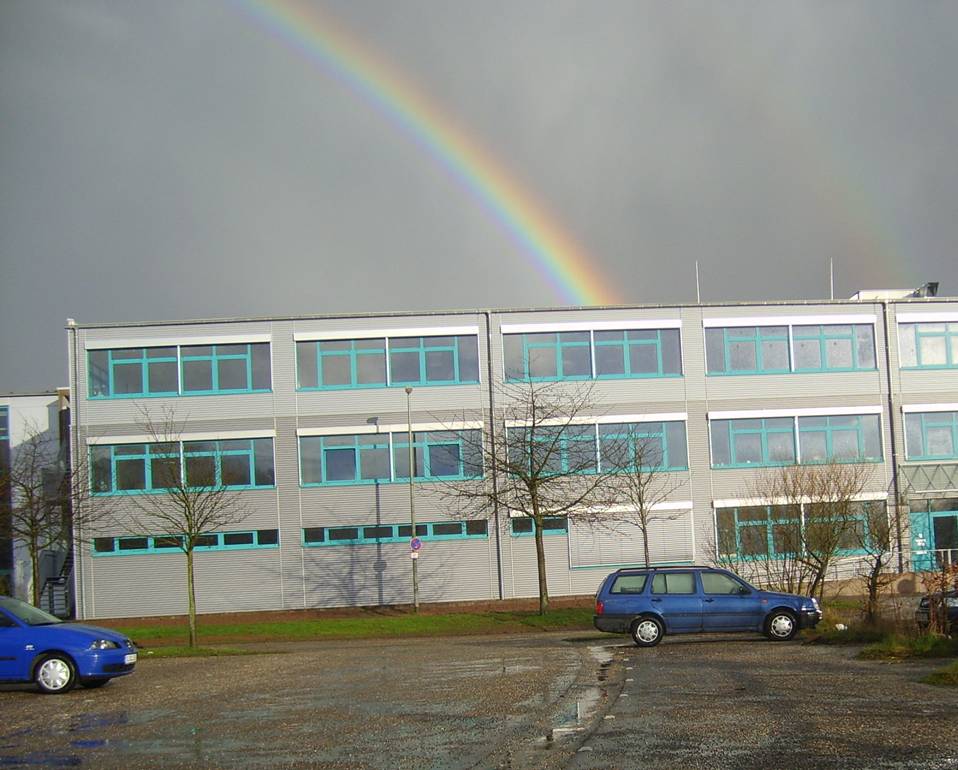 Regenbogen über einem dreistöckigen Bürogebäude mit mehreren Fenstern und zwei geparkten blauen Autos auf einem Parkplatz