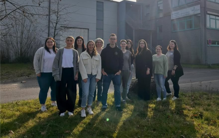 Gruppe von elf Personen steht auf einer Wiese vor einem Gebäude im Hintergrund